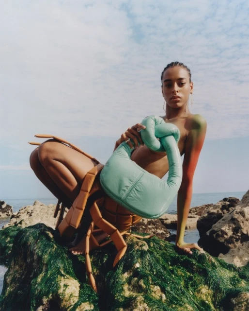 A woman poses on rocks near the ocean holding a handbag, wearing a brown crustacean-inspired garment with orange and green body paint.