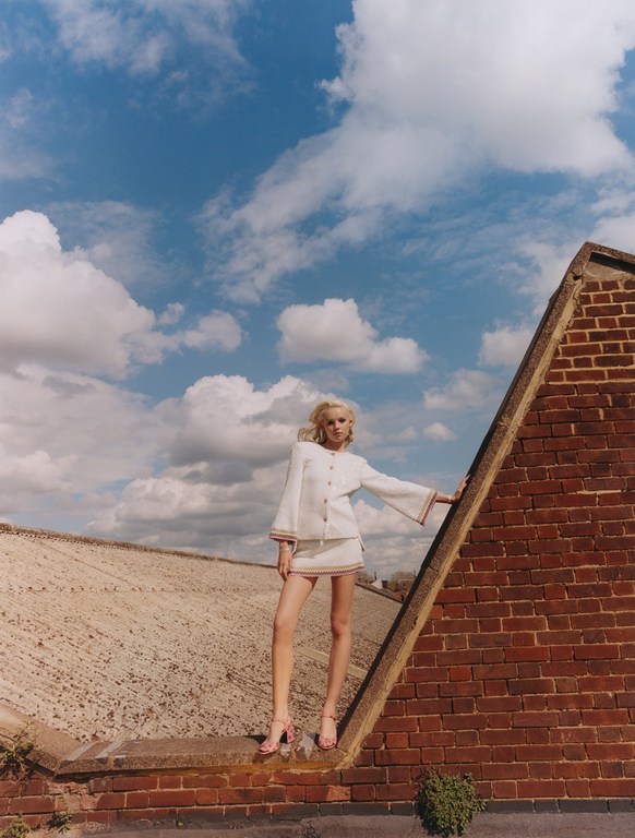 A blonde woman in a white skirt suit poses confidently on a brick rooftop against a backdrop of a partly cloudy sky.