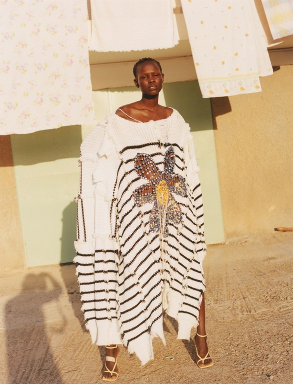 A Black female model wearing a striped sweater dress with embellishments and strappy heels poses outdoors near hanging laundry.
