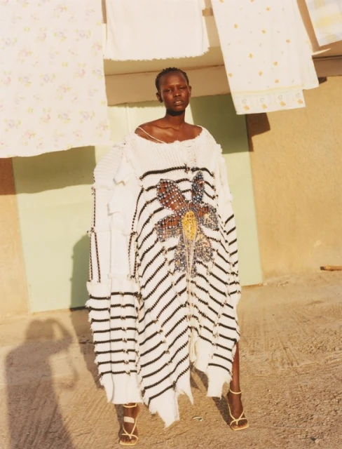 A Black female model wearing a striped sweater dress with embellishments and strappy heels poses outdoors near hanging laundry.