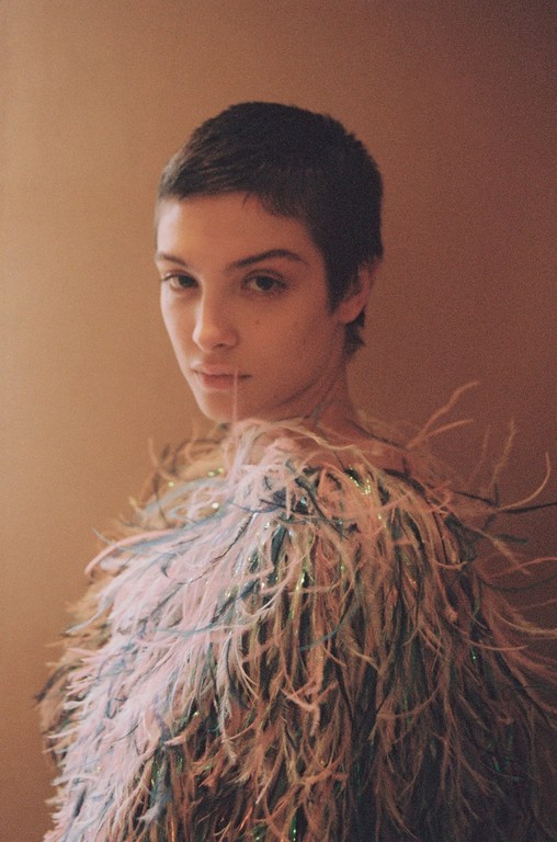Close-up portrait of a model with short hair wearing a pastel feather boa against a neutral tan background in an indoor setting.