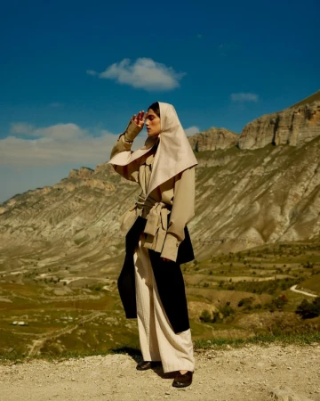 A stylish adult female stands on a hilltop wearing a draped headscarf, trench coat, and wide-leg pants, against a backdrop of mountainous terrain.