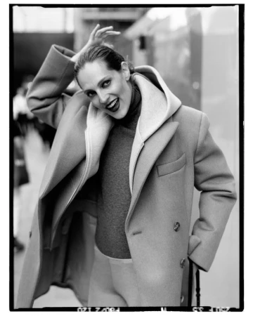 Model in a heavy wool coat with a dark turtleneck smiling with hand raised, captured in monochrome.