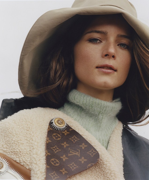 A woman in a cream hat and jacket with a LV-branded patch, facing the camera, showcasing a blend of casual and high fashion elements.