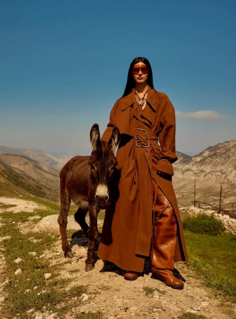 Woman in a brown trench coat poses with a donkey in a sunny outdoor landscape. She's wearing sunglasses and knee-high boots.