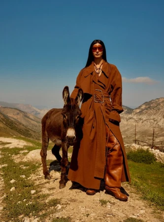 Woman in a brown trench coat poses with a donkey in a sunny outdoor landscape. She's wearing sunglasses and knee-high boots.