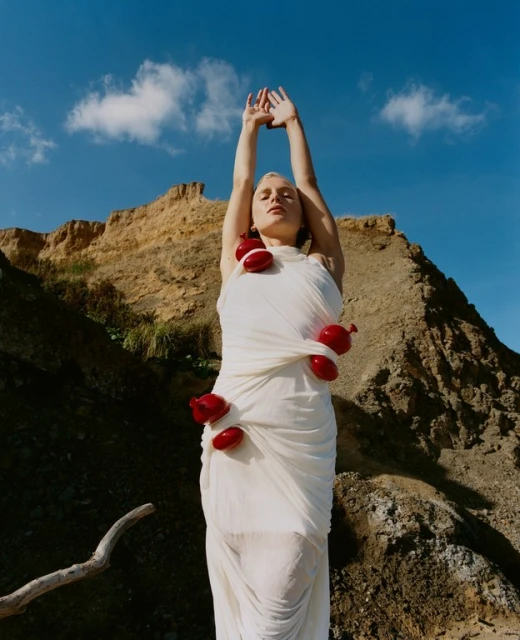 Woman draped in white fabric with red balloons posing against a rugged coastal landscape under a blue sky with clouds.