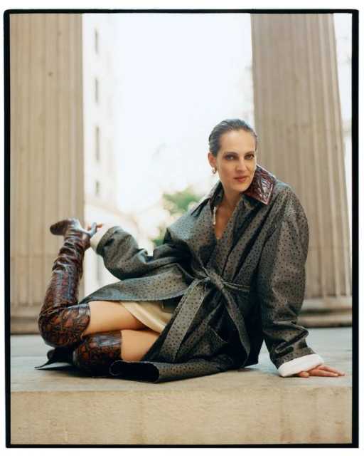 A woman in an open dotted coat and patterned thigh-high boots poses on a stone surface with pillars in the background, fashion shot.