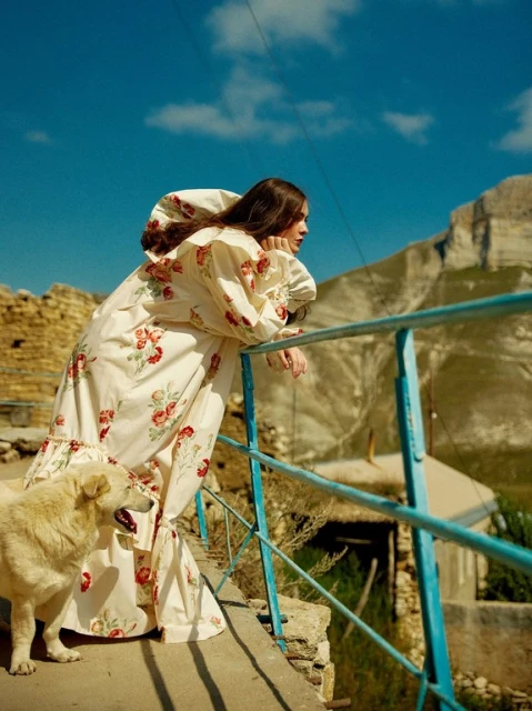 Fashion shot of a woman in a floral dress leaning on a blue railing with a dog in a rural, mountainous outdoor setting.
