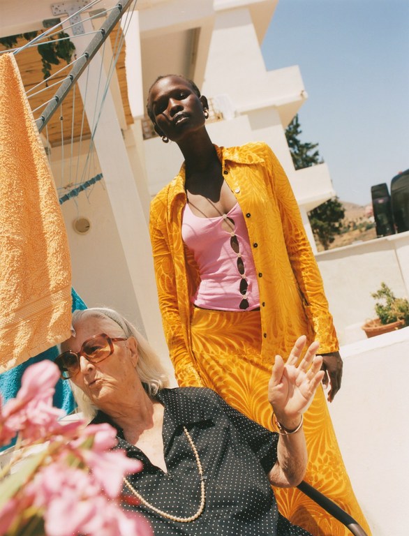 Two women pose on a bright rooftop in the sunshine: one in a polka dot blouse, the other in a pink top with a yellow suit.
