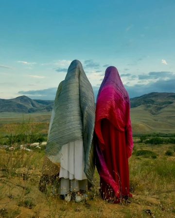 Two people in draped, patterned fabric stand in a grassy landscape with distant mountains beneath a bright blue sky in a wide shot.