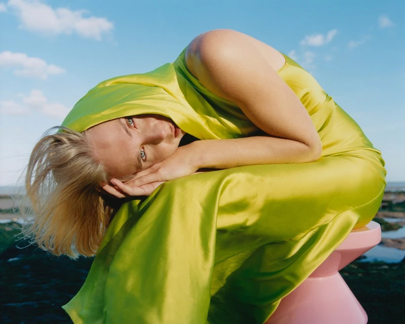 Woman in a chartreuse gown rests on a pink stool outdoors under a blue sky. Her blond hair falls over her face, eyes looking up.