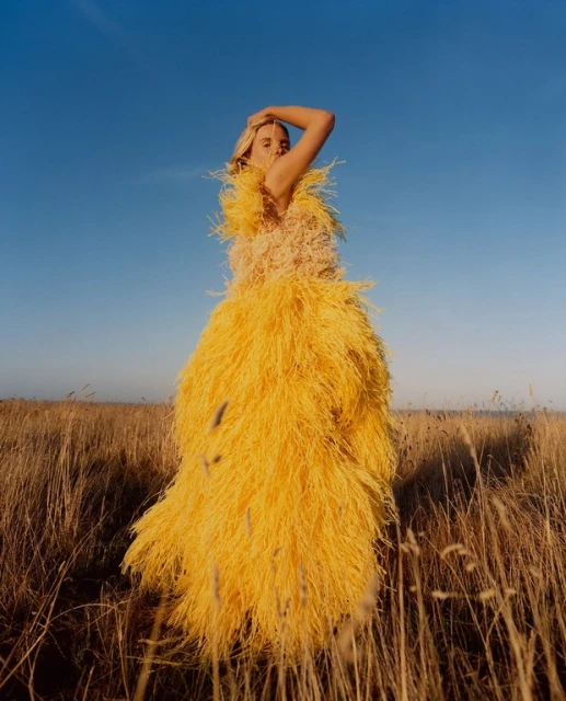 Woman in a yellow, shaggy gown poses in a sunlit field under a clear blue sky for a fashion editorial, capturing a blend of nature and couture.