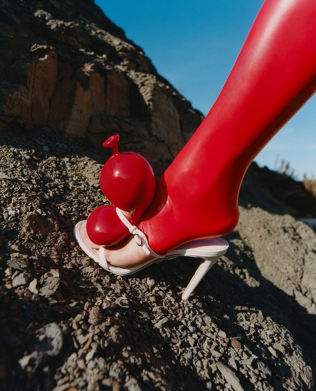 Foot in a high-heeled sandal adorned with two red balloons stands on a rocky surface against a blue sky, in a surreal outdoor fashion shot.