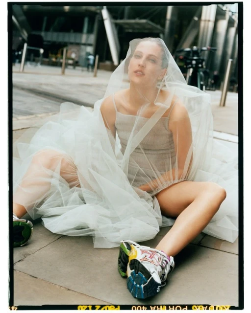 Woman in a tulle dress and sneakers sitting in an urban setting with modern buildings. Light is bright, focus is on the subject.
