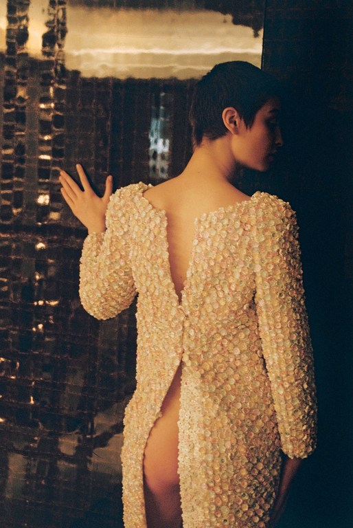 Woman in textured beige jacket, back view, buttons undone, leaning against mirrored wall inside with a dark shadowy ambiance.