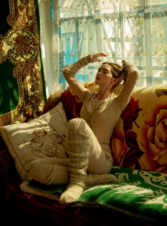 Young woman in a cozy outfit poses with her hands up by a bright window with patterned curtains and textured blankets in a warm indoor setting.