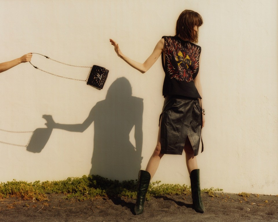 Fashion shot of a woman in a black outfit with a handbag, outdoors against a white wall with plants at the bottom, long shot.
