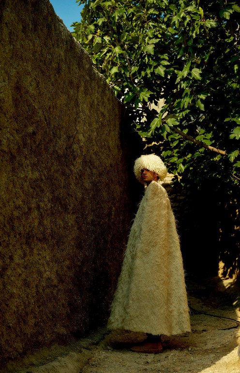 Person in a white, shaggy cloak and hat stands next to a rough wall, framed by green foliage in an outdoor, possibly rural, setting.