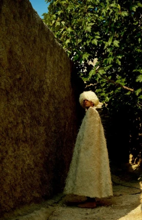 Person in a white, shaggy cloak and hat stands next to a rough wall, framed by green foliage in an outdoor, possibly rural, setting.