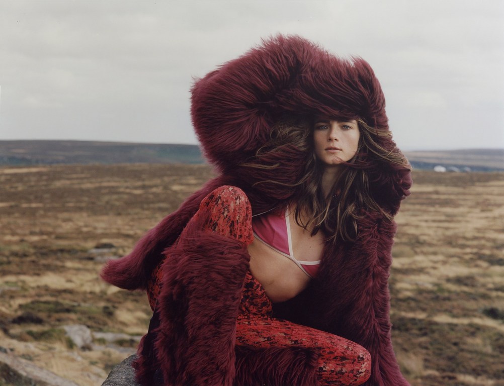 Woman poses in a fur hooded coat and patterned leggings, sitting on a rock outcrop in a wide rural landscape under a cloudy sky.