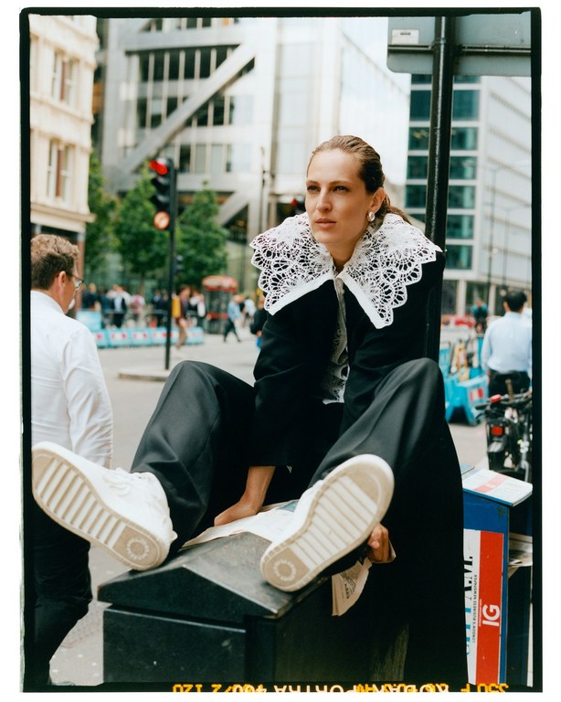 Woman in black suit with lace collar sits on city street in white sneakers with urban backdrop and modern buildings, natural lighting.