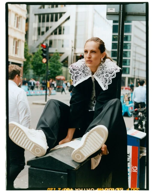 Woman in black suit with lace collar sits on city street in white sneakers with urban backdrop and modern buildings, natural lighting.