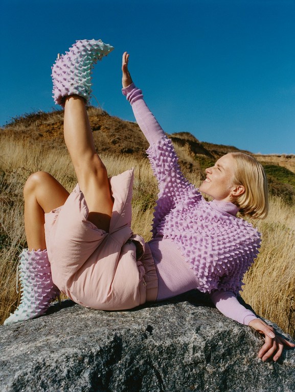 A woman lies on a rock outside, wearing spike-covered boots and a lilac top, one leg raised against a clear blue sky.