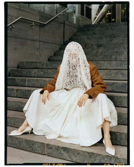 Person in a brown jacket and white skirt sits on grey concrete stairs, face covered with lace veil, wearing white heels and posing.