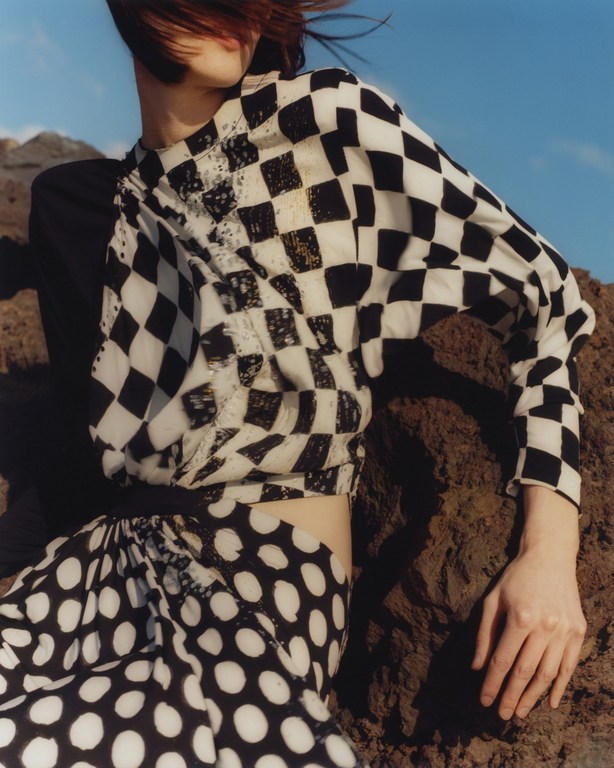 Model in black-and-white checkered top and polka dot skirt poses on sandy terrain against blue sky. Fashion editorial.