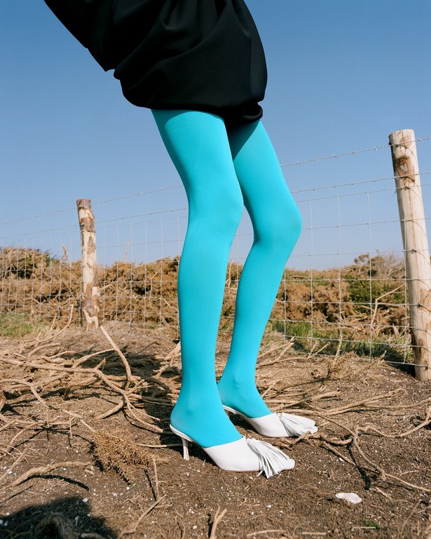 Woman in black dress and bright turquoise tights stands outdoors near a fence and wears white heeled shoes with fringe.