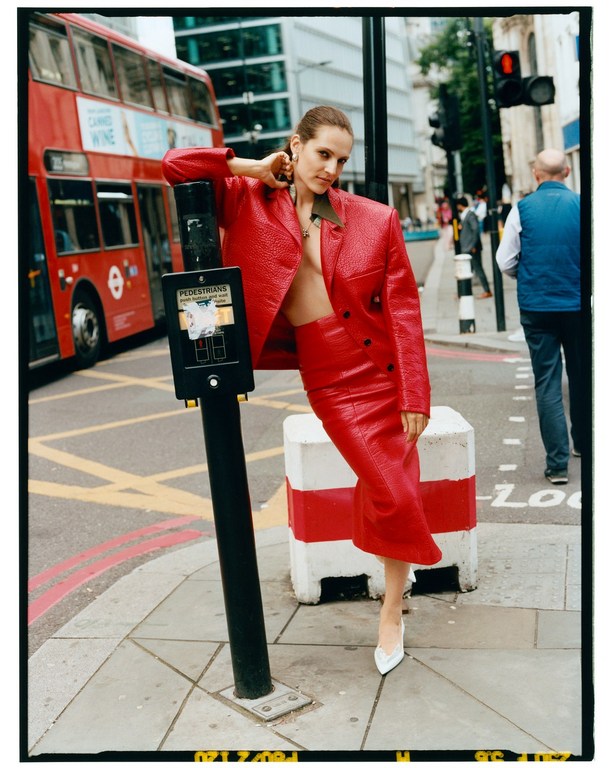 Woman in red leather suit poses on a city sidewalk next to a pedestrian crossing signal. A red double-decker bus drives by.