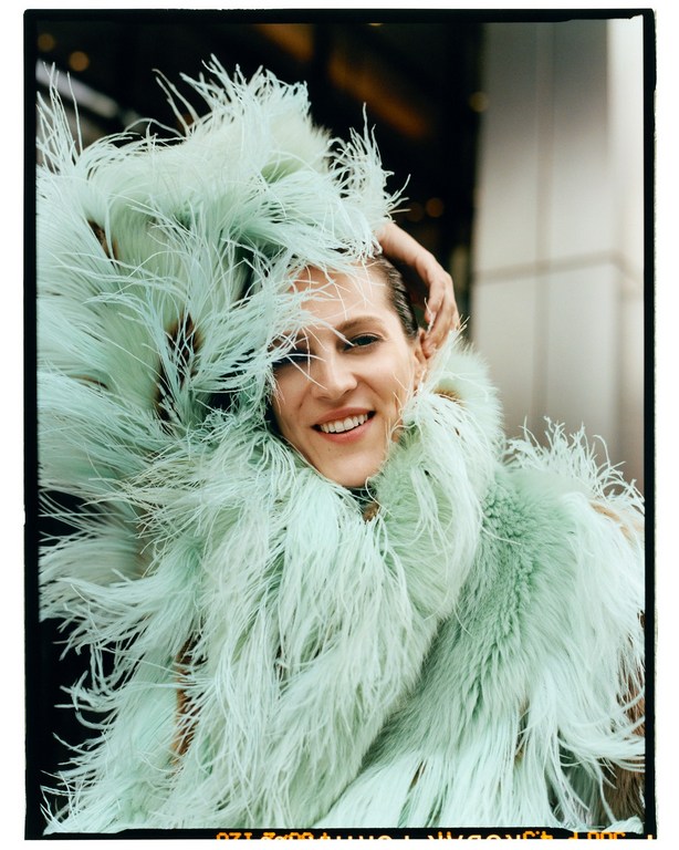 Smiling woman with hand on her head, adorned in light green, feathery boa, standing in an urban setting with a neutral background.