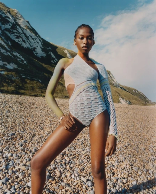 A solo fashion model poses in a knit swimsuit on a pebble beach with a mountain backdrop on a sunny day, looking directly at the camera.