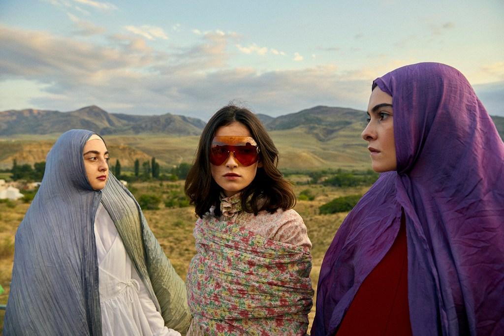 Three women stand outdoors wearing hijabs and dresses; the woman in the middle wears sunglasses and a floral shawl, under a blue sky.