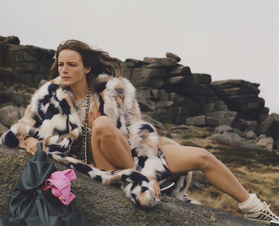 A woman in a colorful fur coat sits on a rock in a grassy, rocky landscape with a backpack, posing for a fashion shot.