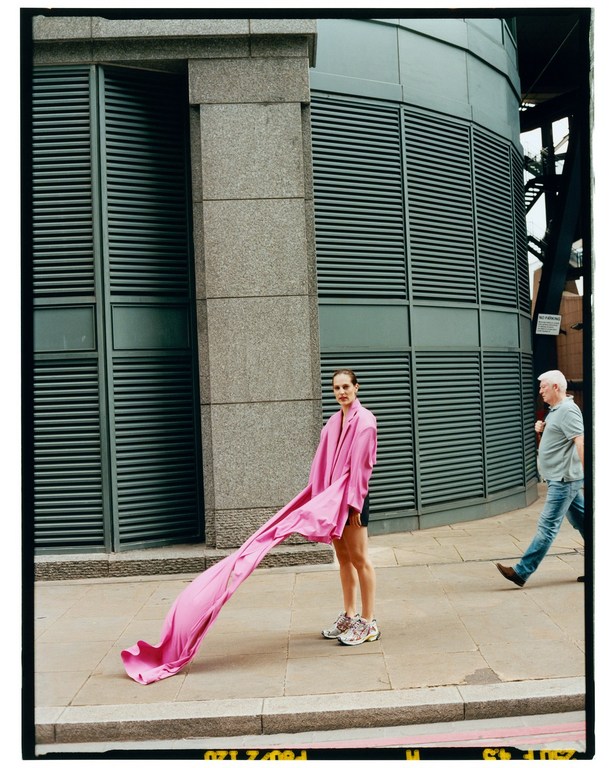 Model standing on a London sidewalk holding a flowing pink garment, with a second person walking past.