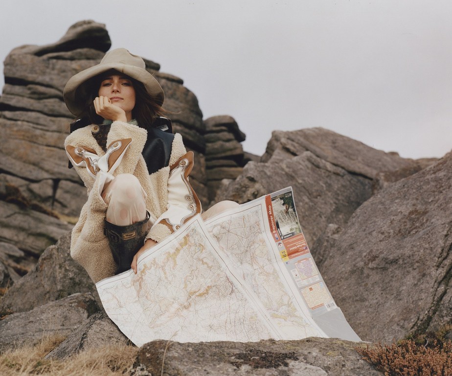 Woman in hat sits among rocks holding a map wearing patterned coat, leggings, and boots in an outdoor, overcast environment.