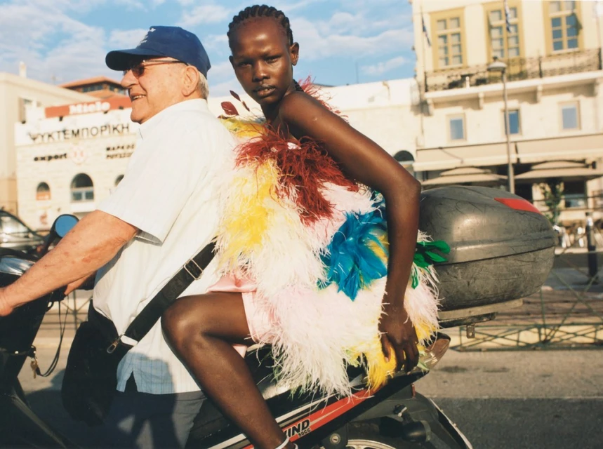 Elderly man drives a motorbike with a young woman wearing a feathered dress in an outdoor urban setting on a sunny day.