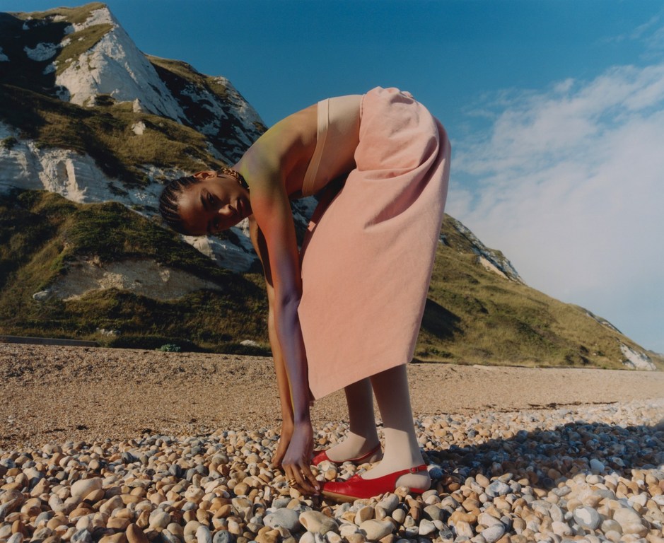 Woman bends at the waist on a pebble beach with chalk cliffs, blue sky. She wears a tan top, pink skirt, red shoes. Lifestyle fashion.