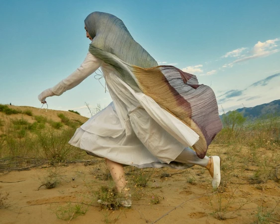Woman in white dress and pleated gradient headscarf running across a sandy desert landscape styled by Tess Yopp.