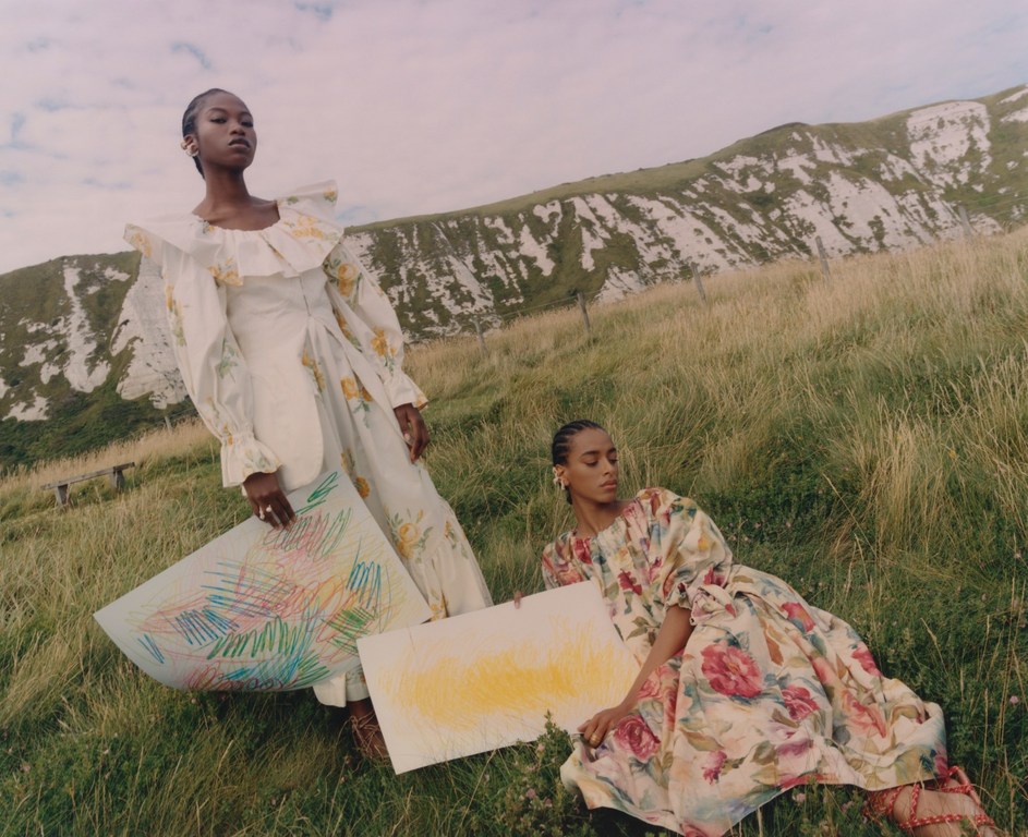 Two dark-skinned women pose in floral dresses in a grassy field with a white chalk hill in the background, holding colorful art pieces.
