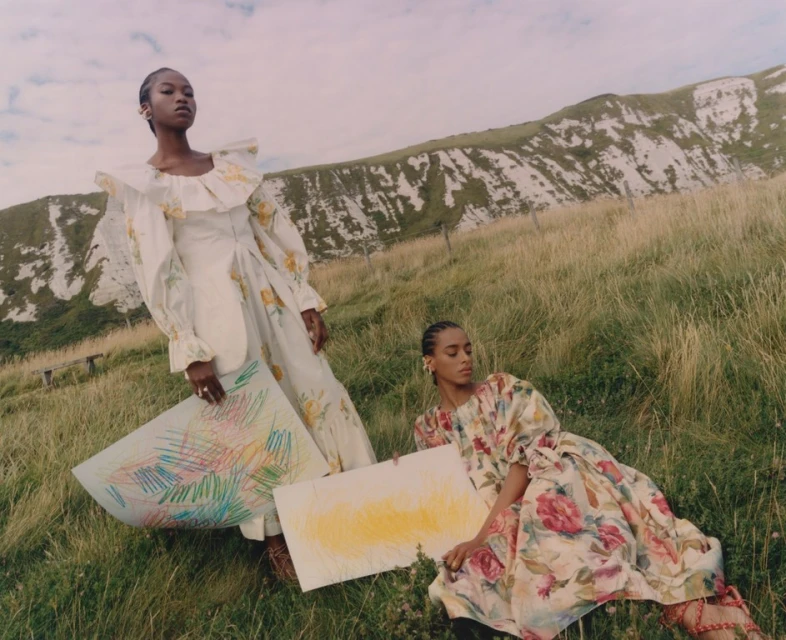 Two dark-skinned women pose in floral dresses in a grassy field with a white chalk hill in the background, holding colorful art pieces.
