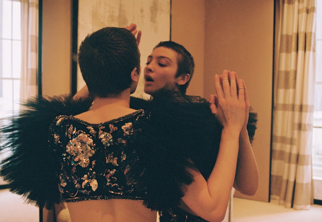 Woman with short hair in embellished black top with feather trim looks at her reflection in a mirror inside of an indoor setting.