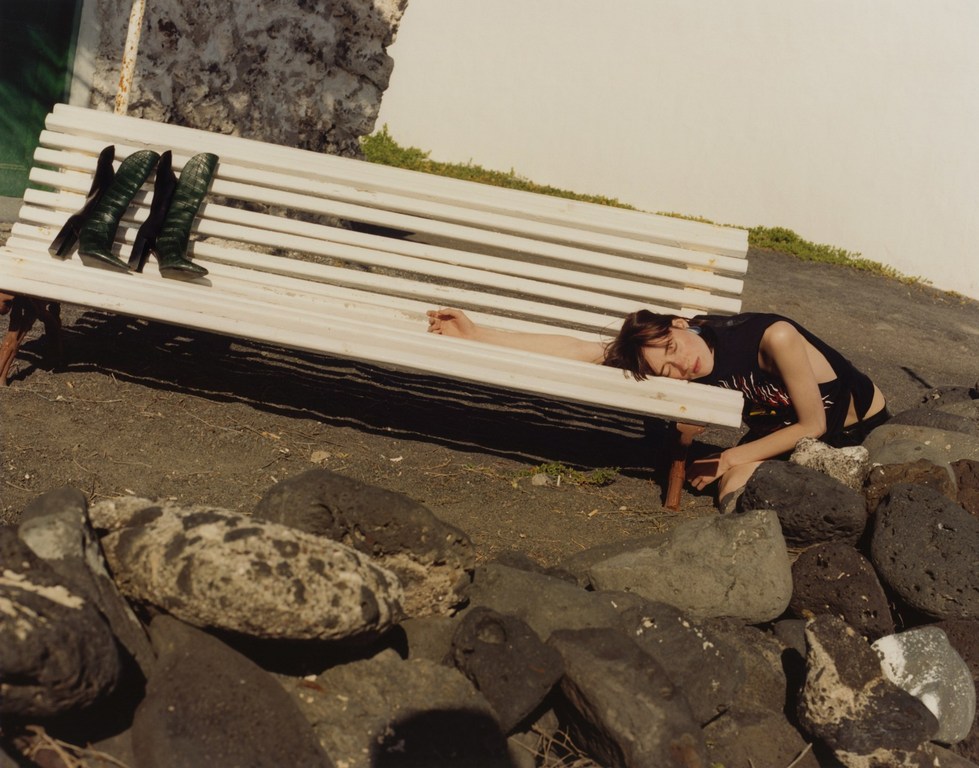 Woman rests on a bench with boots, wearing a tank top, black shorts. A rock wall is in the foreground with an outdoor backdrop.