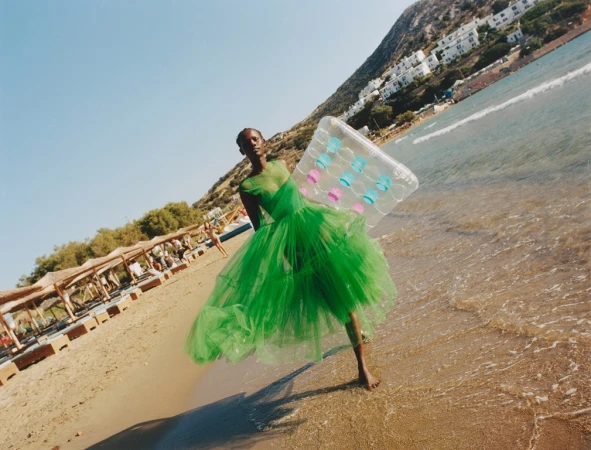 Woman in a green dress walks on a beach holding a clear inflatable raft, with the ocean, beachgoers, and mountains in background.