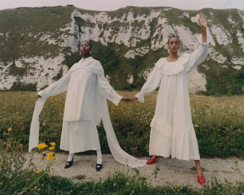 Two women in white dresses holding hands in a grassy field with a large white cliff in the background on a sunny day.