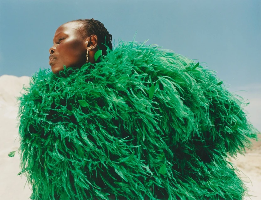 Elegant Black woman poses in an emerald green, feathered, oversized coat against a pale desert and light blue sky in a fashion editorial.