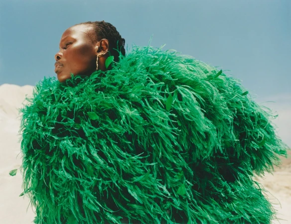 Elegant Black woman poses in an emerald green, feathered, oversized coat against a pale desert and light blue sky in a fashion editorial.