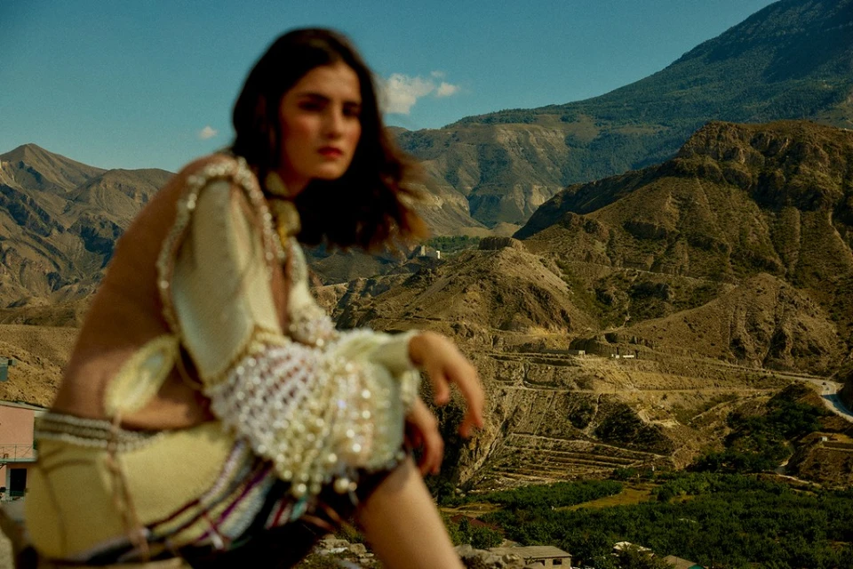 Adult female model poses in a beige embellished outfit in a medium shot against a mountain landscape under a clear sky in daylight.